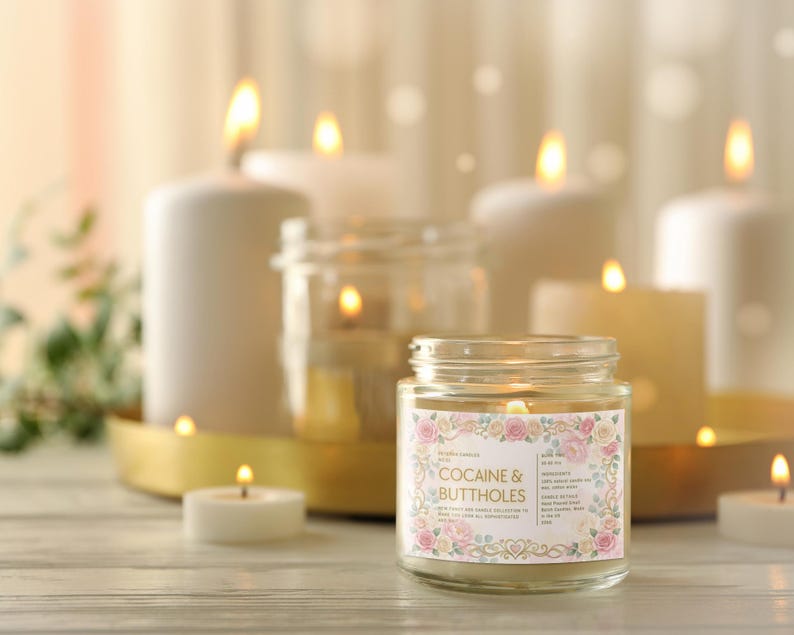 Puede incluir: Una vela encendida en un tarro de cristal con una etiqueta floral que dice "COCAINE & BUTTHOLES" es el punto focal. Varias otras velas encendidas de varios tama&ntilde;os y formas est&aacute;n en el fondo, creando un ambiente c&aacute;lido y acogedor. Las velas est&aacute;n sobre una superficie de madera.