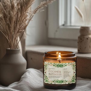 Puede incluir: Una vela encendida en un tarro de vidrio &aacute;mbar con una etiqueta que dice "Cocaine & Buttholes". La vela est&aacute; rodeada por un dise&ntilde;o floral verde. La vela est&aacute; sobre una tela blanca con un jarr&oacute;n de plantas secas en el fondo.