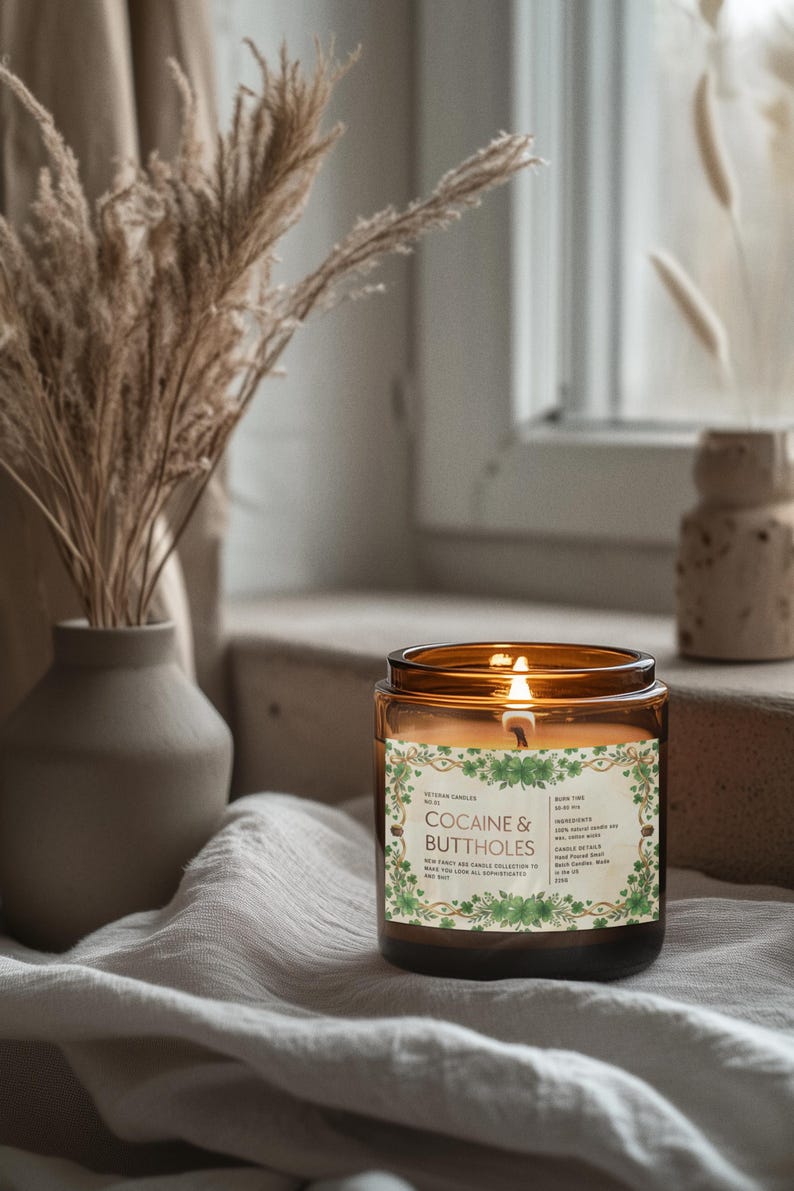 Puede incluir: Una vela encendida en un tarro de vidrio &aacute;mbar con una etiqueta que dice "Cocaine & Buttholes". La vela est&aacute; rodeada por un dise&ntilde;o floral verde. La vela est&aacute; sobre una tela blanca con un jarr&oacute;n de plantas secas en el fondo.