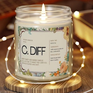 May include: A lit glass jar candle with a label that reads "C. DIFF" and "New Fancy Ass Nurse Candle Collection". The candle has floral accents and is on a wooden coaster, surrounded by fairy lights. The candle's burn time is 45-50 hours.