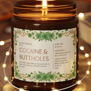 Puede incluir: Una vela encendida en un tarro de cristal marr&oacute;n con una etiqueta que dice "COCAINE & BUTTHOLES". La etiqueta tiene un dise&ntilde;o de tr&eacute;bol verde y el texto dice "New Fancy Ass Candle Collection to Make You Look All Sophisticated and Shit". La vela est&aacute; sobre un posavasos de madera.