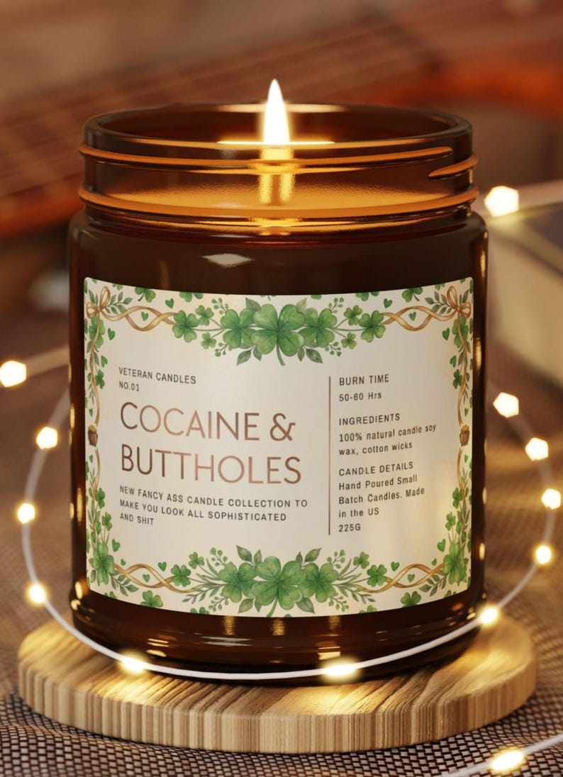 Puede incluir: Una vela encendida en un tarro de cristal marr&oacute;n con una etiqueta que dice "COCAINE & BUTTHOLES". La etiqueta tiene un dise&ntilde;o de tr&eacute;bol verde y el texto dice "New Fancy Ass Candle Collection to Make You Look All Sophisticated and Shit". La vela est&aacute; sobre un posavasos de madera.