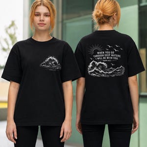 May include: A black t-shirt featuring a white graphic of a crashing wave on the front chest with the text Isaiah 43:2. The back displays a larger white graphic of a wave, a sun, and birds with the text WHEN YOU GO THROUGH DEEP WATERS, I WILL BE WITH YOU. ISAIAH 43:2.