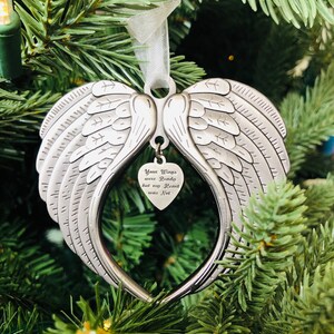 May include: A silver angel wing ornament with a heart-shaped charm that reads "Your Wings were Ready but my Heart was Not". The ornament is hanging on a green Christmas tree.