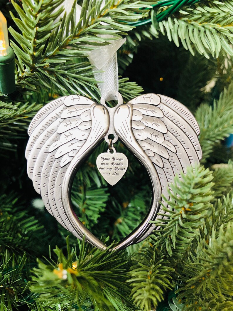 May include: A silver angel wing ornament with a heart-shaped charm that reads "Your Wings were Ready but my Heart was Not". The ornament is hanging on a green Christmas tree.