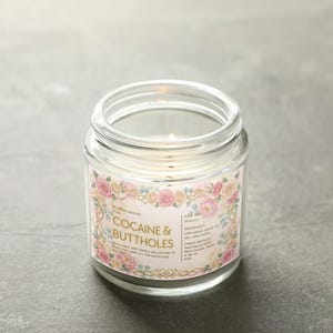 Puede incluir: Una vela en tarro de cristal transparente con una mecha encendida. La etiqueta dice "Cocaine & Buttholes" en una fuente decorativa, rodeada de un dise&ntilde;o floral con rosas rosas y crema. La vela est&aacute; sobre una superficie gris.