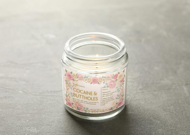 Puede incluir: Una vela en tarro de cristal transparente con una mecha encendida. La etiqueta dice "Cocaine & Buttholes" en una fuente decorativa, rodeada de un dise&ntilde;o floral con rosas rosas y crema. La vela est&aacute; sobre una superficie gris.