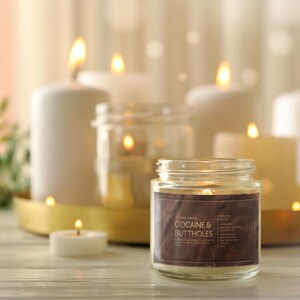 May include: A lit candle in a glass jar with a brown label that reads "COCAINE & BUTTHOLES" sits on a wooden surface. Several other lit candles of varying sizes and shapes are in the background, creating a warm, inviting ambiance.