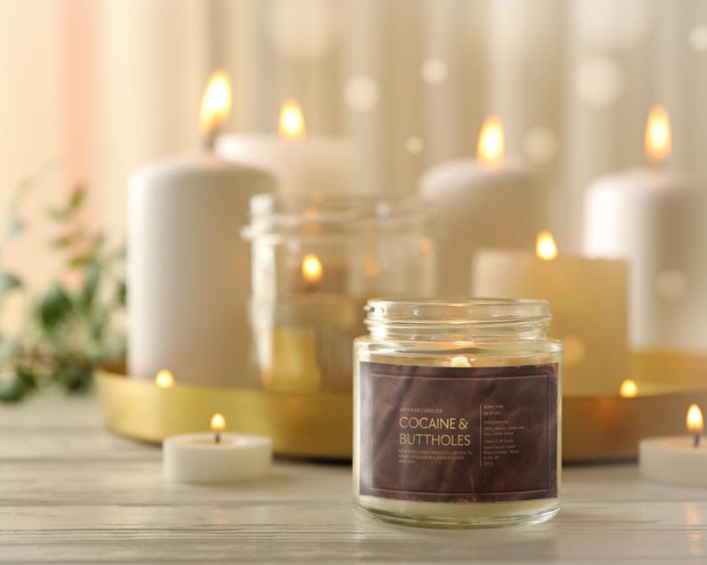 May include: A lit candle in a glass jar with a brown label that reads "COCAINE & BUTTHOLES" sits on a wooden surface. Several other lit candles of varying sizes and shapes are in the background, creating a warm, inviting ambiance.