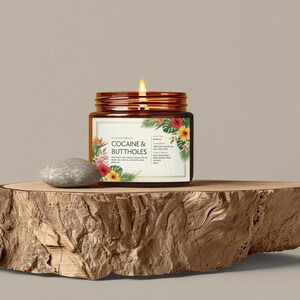 Może przedstawiać: Zapalone świeca w brązowym szklanym słoiku z etykietą z napisem "COCAINE & BUTTHOLES" z kwiatowymi akcentami. Słoik stoi na drewnianej płycie obok szarego kamienia. Tło jest w jednolitym neutralnym kolorze.