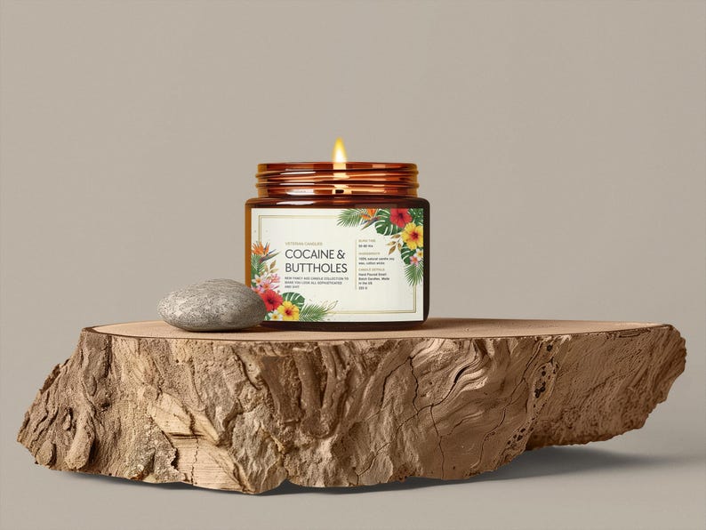 Może przedstawiać: Zapalone świeca w brązowym szklanym słoiku z etykietą z napisem "COCAINE & BUTTHOLES" z kwiatowymi akcentami. Słoik stoi na drewnianej płycie obok szarego kamienia. Tło jest w jednolitym neutralnym kolorze.