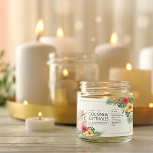 May include: A lit jar candle with a floral label that reads "COCAINE & BUTTHOLES" is the focal point, surrounded by other lit candles of varying sizes. The candles are on a gold tray and a wooden surface.
