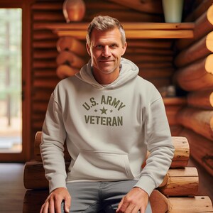 Puede incluir: Una sudadera con capucha gris claro con el texto "U.S. ARMY VETERAN" en verde oliva. La sudadera tiene un bolsillo delantero y una capucha. La persona lleva pantalones grises y est&aacute; sentada en un banco de madera en una caba&ntilde;a de troncos.