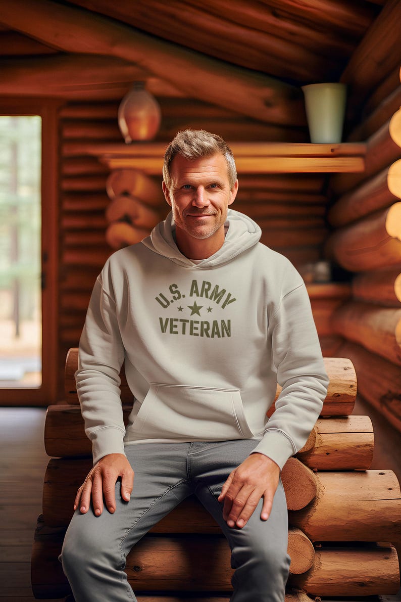 Puede incluir: Una sudadera con capucha gris claro con el texto "U.S. ARMY VETERAN" en verde oliva. La sudadera tiene un bolsillo delantero y una capucha. La persona lleva pantalones grises y est&aacute; sentada en un banco de madera en una caba&ntilde;a de troncos.
