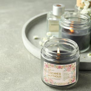 Puede incluir: Una vela gris oscura encendida en un tarro de cristal transparente con una etiqueta floral que dice "Cocaine & Buttholes". La vela est&aacute; sobre una bandeja gris con otras velas y peque&ntilde;as flores blancas.