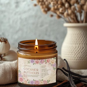 May include: A lit brown glass jar candle with a label that reads "COCAINE & BUTTHOLES" surrounded by floral designs. The candle sits on a wooden surface with cinnamon sticks and vanilla beans. A vase with dried flowers is in the background.