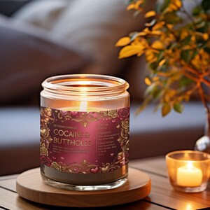 Peut inclure: Une bougie allum&eacute;e dans un bocal en verre avec une &eacute;tiquette qui dit "COCAINE & BUTTHOLES". L'&eacute;tiquette est bordeaux avec des accents dor&eacute;s et des illustrations de roses. La bougie repose sur un dessous de verre en bois, avec une petite bougie votive &agrave; droite.