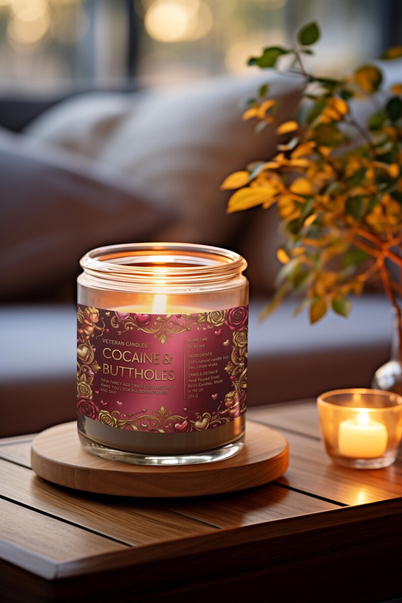 Peut inclure: Une bougie allum&eacute;e dans un bocal en verre avec une &eacute;tiquette qui dit "COCAINE & BUTTHOLES". L'&eacute;tiquette est bordeaux avec des accents dor&eacute;s et des illustrations de roses. La bougie repose sur un dessous de verre en bois, avec une petite bougie votive &agrave; droite.