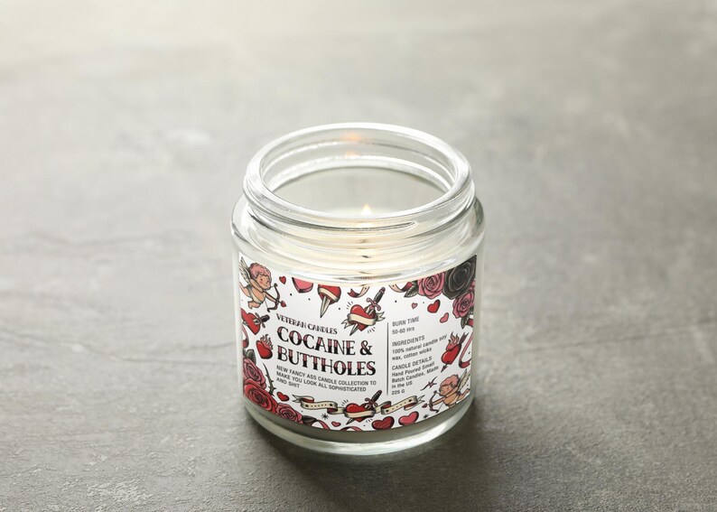 May include: A lit, clear glass jar candle with a label that reads "Cocaine & Buttholes." The label features tattoo-style illustrations of hearts, roses, and cherubs. The candle is part of the "New Fancy Ass Candle Collection."