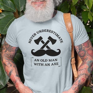 May include: Light blue t-shirt with the text "NEVER UNDERESTIMATE AN OLD MAN WITH AN AXE." The design includes crossed axes and a mustache. The shirt is worn by a person with tattoos and a beard.