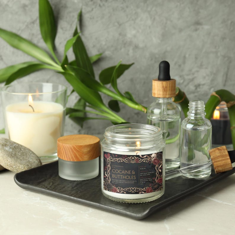 Pu&ograve; includere: Una candela accesa in un barattolo di vetro con l'etichetta "COCAINE & BUTTHOLES" &egrave; posta su un vassoio nero, insieme ad altre candele, bottiglie di vetro e bamb&ugrave;. L'etichetta ha un bordo decorativo con teschi e rose.