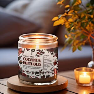 Może przedstawiać: Zapalone świeca w szklanym słoiku z napisem "COCAINE & BUTTHOLES" na etykiecie. Etykieta zawiera ilustracje w stylu tatuażu. Świeca stoi na drewnianej podstawce, w pobliżu mała zapalona świeca.