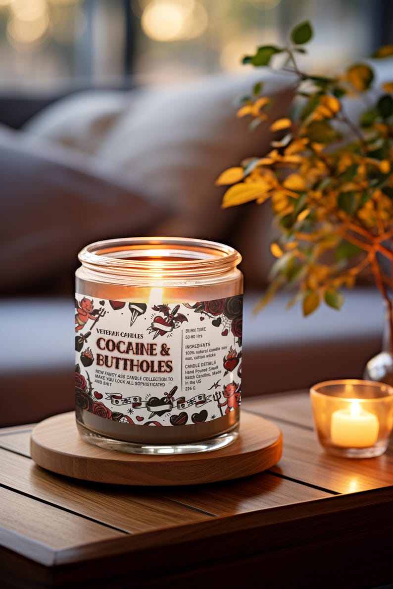 Może przedstawiać: Zapalone świeca w szklanym słoiku z napisem "COCAINE & BUTTHOLES" na etykiecie. Etykieta zawiera ilustracje w stylu tatuażu. Świeca stoi na drewnianej podstawce, w pobliżu mała zapalona świeca.