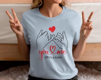 Puede incluir: Camiseta azul claro con un gráfico de dos manos haciendo un juramento de meñique, un corazón rojo y las palabras "you & me Forever & Always" en escritura roja. La camiseta es de cuello redondo.