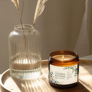 May include: A lit candle in a brown glass jar with a label that reads "COCAINE & BUTTHOLES" sits on a wooden tray. A clear glass vase with decorative dried grass is next to the candle. The candle has a white label with blue floral accents.