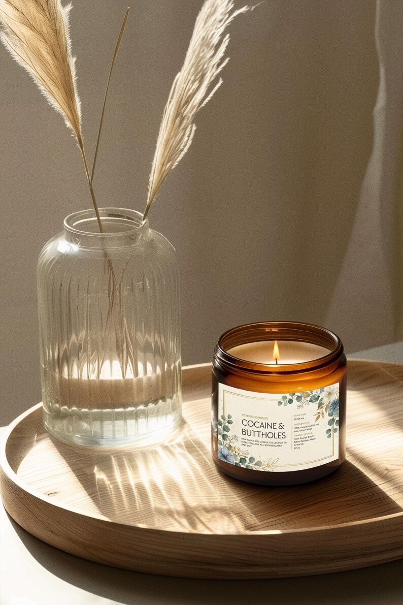 May include: A lit candle in a brown glass jar with a label that reads "COCAINE & BUTTHOLES" sits on a wooden tray. A clear glass vase with decorative dried grass is next to the candle. The candle has a white label with blue floral accents.