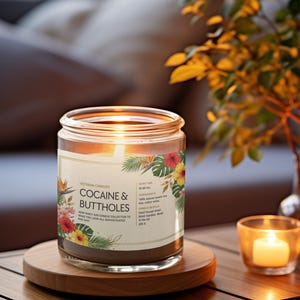 May include: A lit candle in a glass jar with a label that reads "COCAINE & BUTTHOLES." The candle sits on a wooden coaster, with a smaller candle and foliage in the background. The label has floral accents.