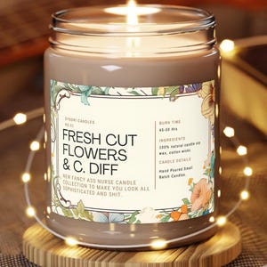 May include: A brown candle in a glass jar with a floral label. The label reads "FRESH CUT FLOWERS & C. DIFF" and "NEW FANCY ASS NURSE CANDLE COLLECTION". The candle is lit and sits on a wooden coaster, with fairy lights around it.