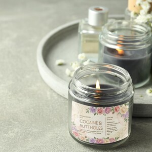 May include: A lit, dark gray candle in a clear glass jar with a floral label that reads "COCAINE & BUTTHOLES." The candle sits on a gray tray with other candles and small white flowers. The background is a light gray surface.