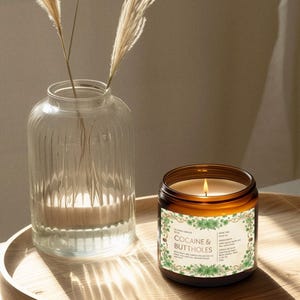 Puede incluir: Una vela encendida en un tarro de cristal marr&oacute;n con una etiqueta que dice "COCAINE & BUTTHOLES" est&aacute; sobre una bandeja de madera. A la izquierda hay un jarr&oacute;n de cristal transparente con plantas secas. La llama de la vela proyecta un brillo c&aacute;lido.