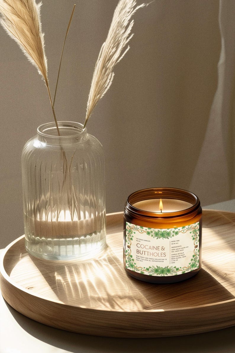 Puede incluir: Una vela encendida en un tarro de cristal marr&oacute;n con una etiqueta que dice "COCAINE & BUTTHOLES" est&aacute; sobre una bandeja de madera. A la izquierda hay un jarr&oacute;n de cristal transparente con plantas secas. La llama de la vela proyecta un brillo c&aacute;lido.