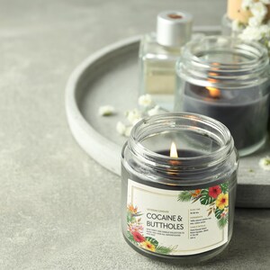 May include: A lit, dark grey candle in a clear glass jar with a label featuring floral designs and the text "Cocaine & Buttholes." Other candles and a perfume bottle are in the background on a grey tray.