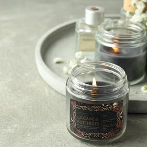 Pu&ograve; includere: Una candela grigio scuro accesa in un barattolo di vetro trasparente con un'etichetta che recita "COCAINE & BUTTHOLES". L'etichetta ha un bordo decorativo con teschi e rose. Altre candele e bottiglie sono sullo sfondo.