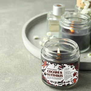 Może przedstawiać: Zapalone, przezroczyste świece w słoiku z czarnym woskiem w środku. Etykieta głosi "Cocaine & Buttholes" z grafiką w stylu tatuażu. Inne świece i taca znajdują się w tle.