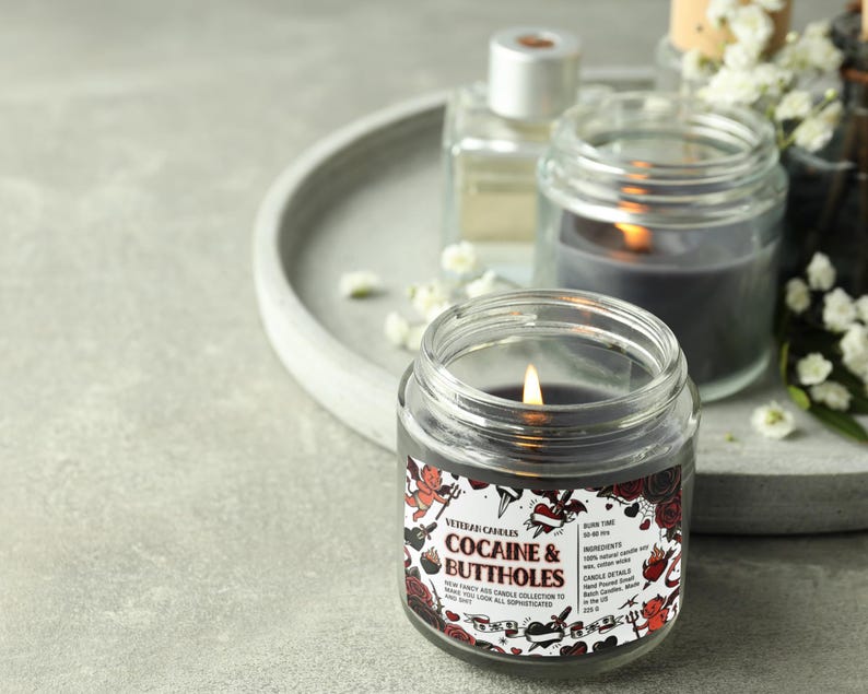 Może przedstawiać: Zapalone, przezroczyste świece w słoiku z czarnym woskiem w środku. Etykieta głosi "Cocaine & Buttholes" z grafiką w stylu tatuażu. Inne świece i taca znajdują się w tle.