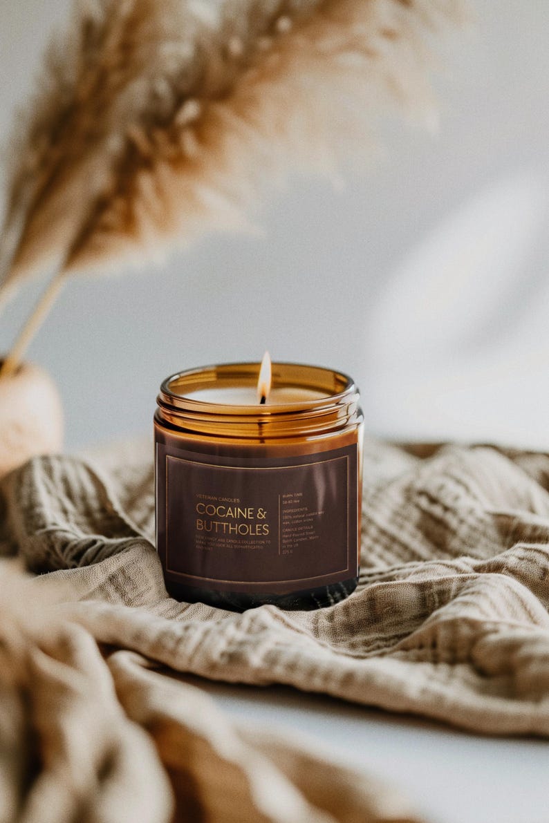 May include: A lit candle in a clear glass jar with a brown label that reads "COCAINE & BUTTHOLES". The candle sits on a beige cloth. The background includes dried plants.