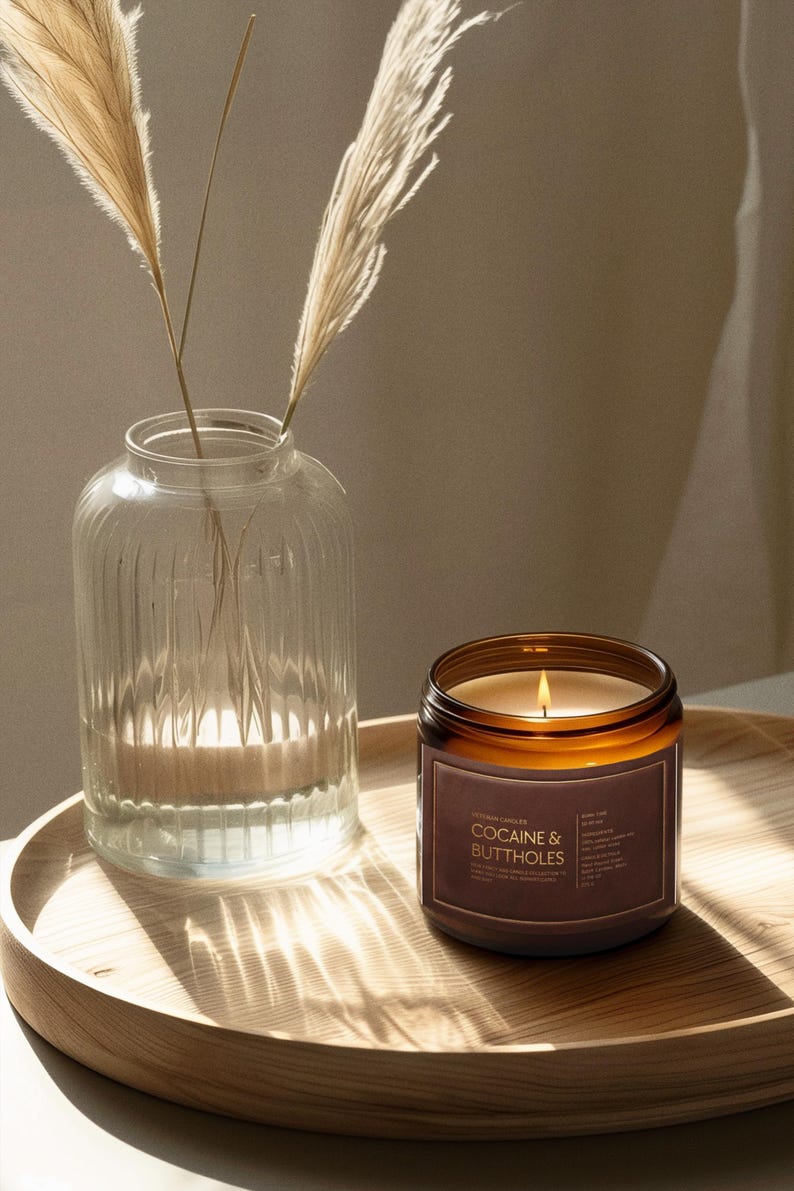 May include: A lit candle in a brown glass jar with a label that reads "COCAINE & BUTTHOLES" sits on a wooden tray. A clear glass vase with dried ornamental grass is to the left. The background is a neutral color.