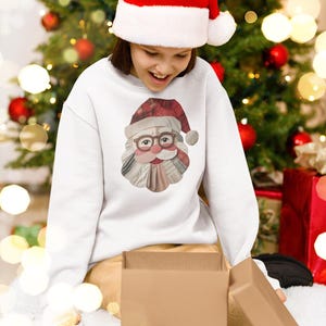 May include: A person wearing a white sweatshirt with a Santa Claus graphic and a red and white Santa hat. The person is opening a brown cardboard box. A Christmas tree with red baubles is in the background.