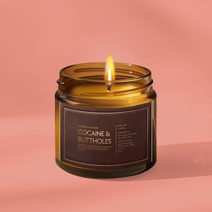 May include: A lit candle in a brown glass jar with a label that reads "COCAINE & BUTTHOLES" in white text. The candle has a bright yellow flame and sits on a pink background.