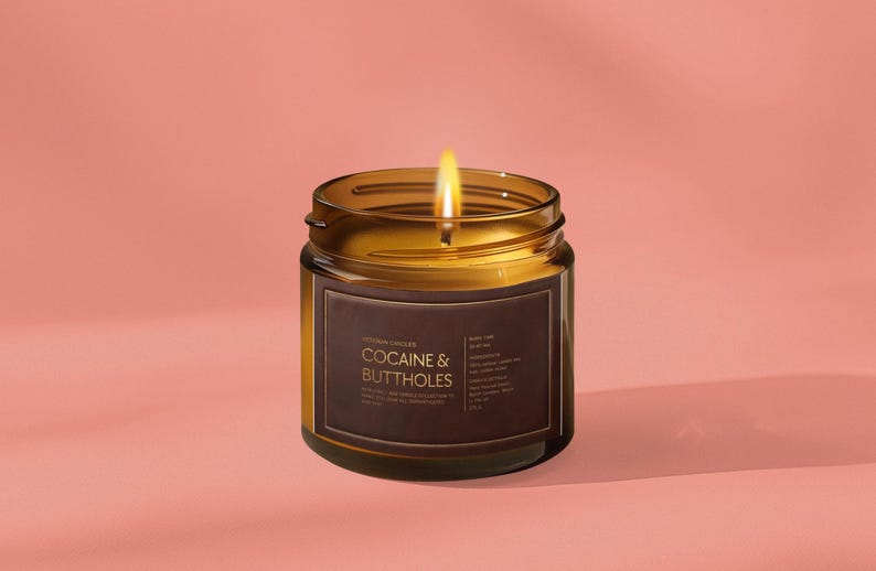May include: A lit candle in a brown glass jar with a label that reads "COCAINE & BUTTHOLES" in white text. The candle has a bright yellow flame and sits on a pink background.