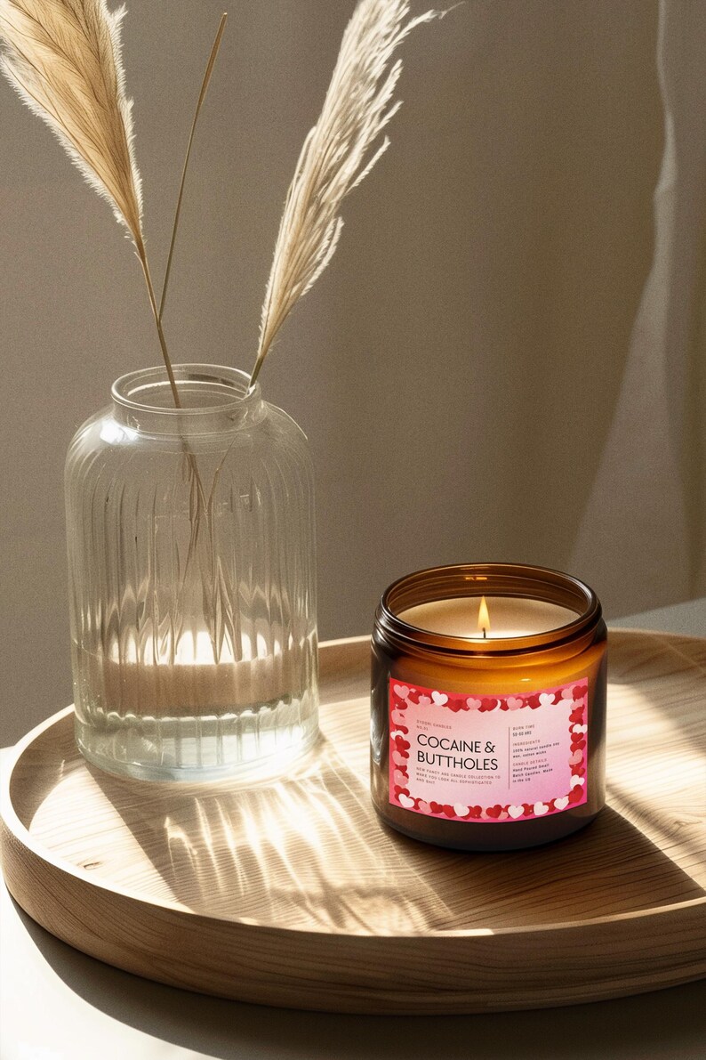 May include: A lit candle in a brown glass jar with a label that reads "COCAINE & BUTTHOLES" sits on a wooden tray. A clear glass vase with dried plants is to the left of the candle. The background is a neutral color.