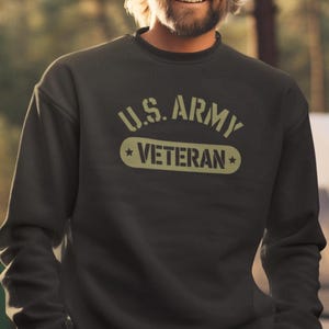May include: Dark olive green sweatshirt with the text "U.S. ARMY VETERAN" in a light olive green color. The sweatshirt has a crew neck and long sleeves. The image shows a person wearing the sweatshirt.