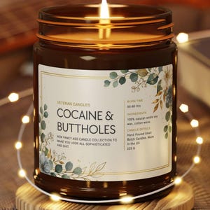 May include: A lit brown glass jar candle with a label that reads "COCAINE & BUTTHOLES". The label has floral accents and the text "Veteran Candles". The candle sits on a wooden coaster and is surrounded by string lights.
