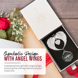 May include: A silver angel wing ornament with a heart pendant and a faceted crystal. The ornament is displayed in a white gift box with the text "Symbolic Design WITH ANGEL WINGS". A red rose and white flowers are in the background.