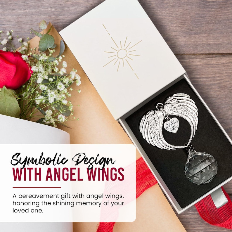 May include: A silver angel wing ornament with a heart pendant and a faceted crystal. The ornament is displayed in a white gift box with the text "Symbolic Design WITH ANGEL WINGS". A red rose and white flowers are in the background.