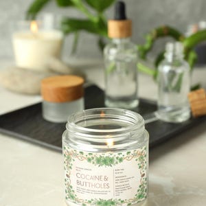 Op de afbeelding: Een brandende glazen potkaars met een label met de tekst "Cocaine & Buttholes" omgeven door een groen bloemmotief. De kaars maakt deel uit van de Veteran Candles-collectie. Andere glazen flessen en een pot met houten deksel staan op de achtergrond.